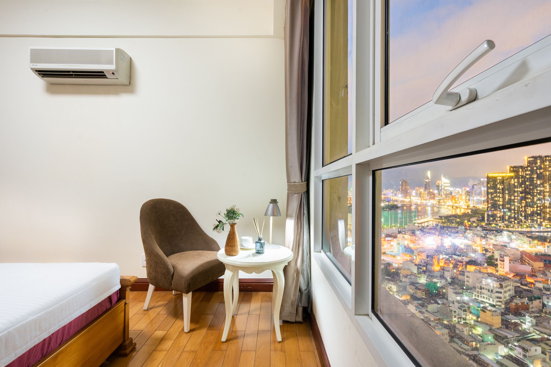 Habitación de hotel con una cama, una silla, una mesita con un jarrón y una lámpara, y un ventanal con vistas a la ciudad.