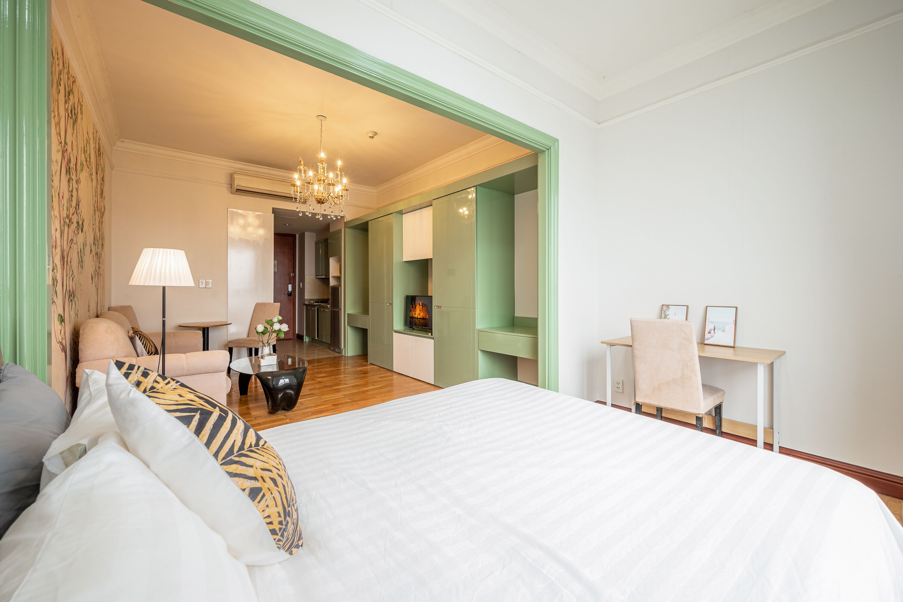 A hotel room with a large bed, a desk, a chair, and a small table with a lamp. There is a fireplace in the background.