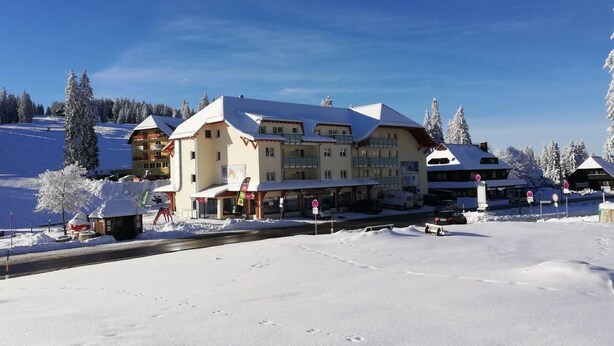 Un hotel grande con varios pisos, un telesilla y árboles nevados al fondo.
