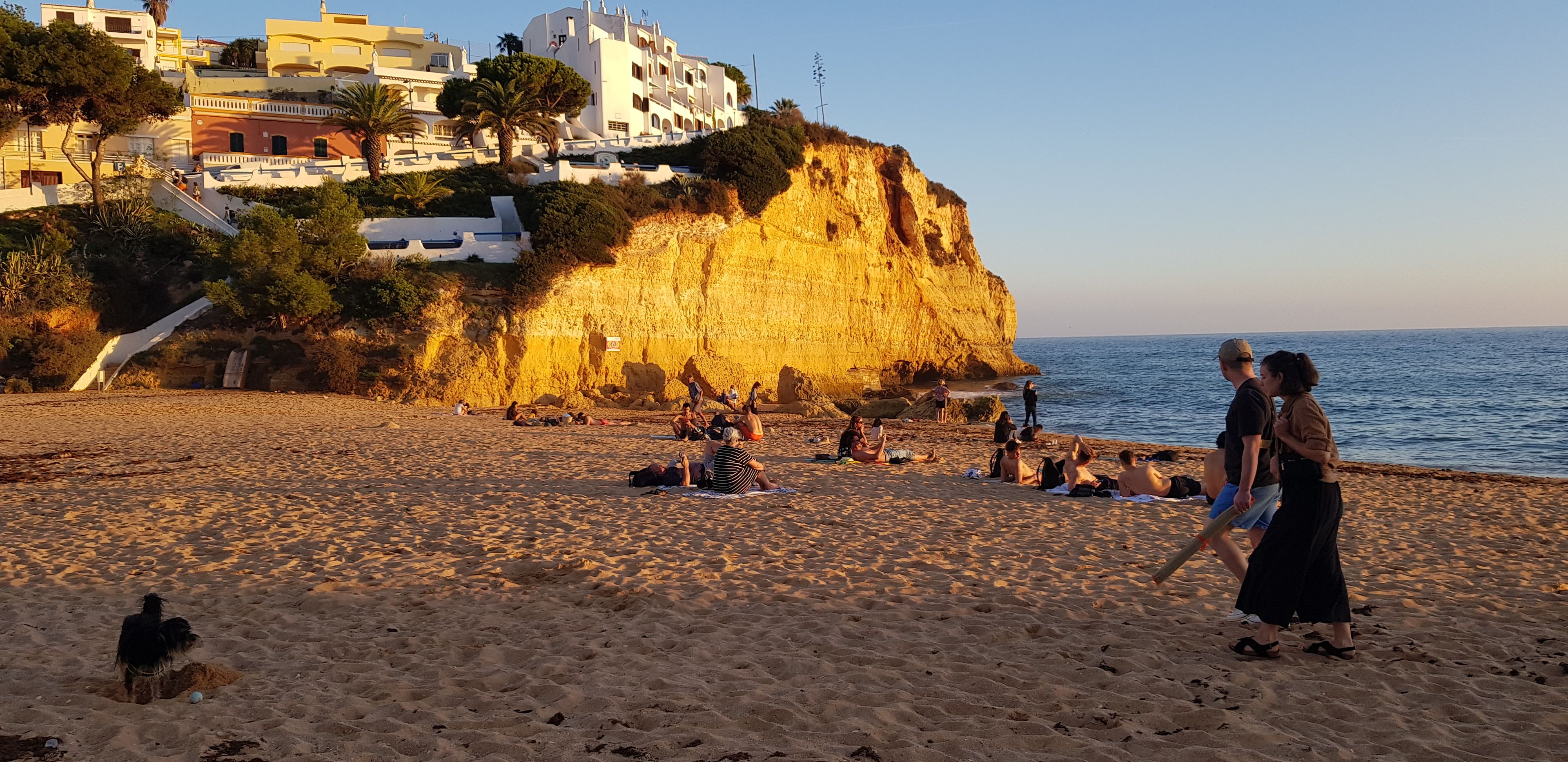 Una playa con gente tomando el sol y caminando, un acantilado con edificios, y el mar al fondo.