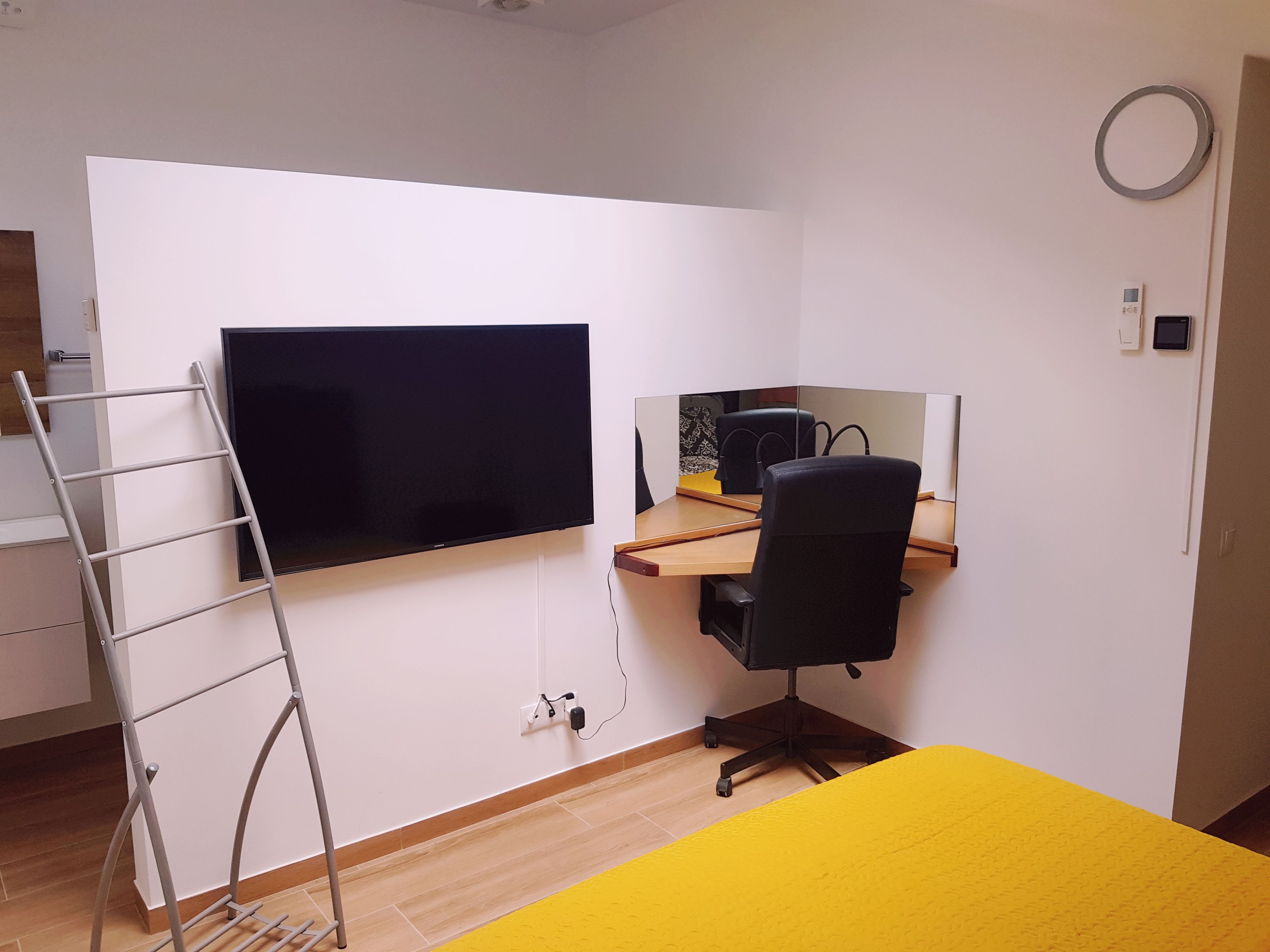 Une chambre dotée d’un téléviseur à écran plat fixé au mur, d’un bureau avec un ordinateur et une chaise, et d’un lit jaune.