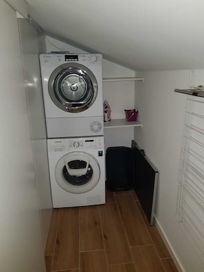 A laundry room with two stacked washing machines, a shelf with a pink item, and a black trash bin.