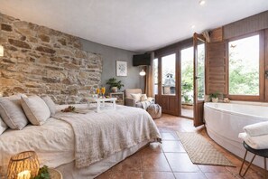 A bedroom with a stone wall, a bed, a bedside table with glasses, a chair, and a bathtub.