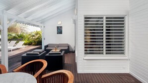 Un espace extérieur abrité, doté d’une façade blanche, d’une grande fenêtre avec des stores et d’un coin salon comprenant un canapé et des fauteuils.
