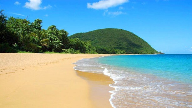 Ein Strand mit klarem türkisblauem Wasser, einem sandigen Ufer und einer üppigen grünen Hügelkette im Hintergrund.