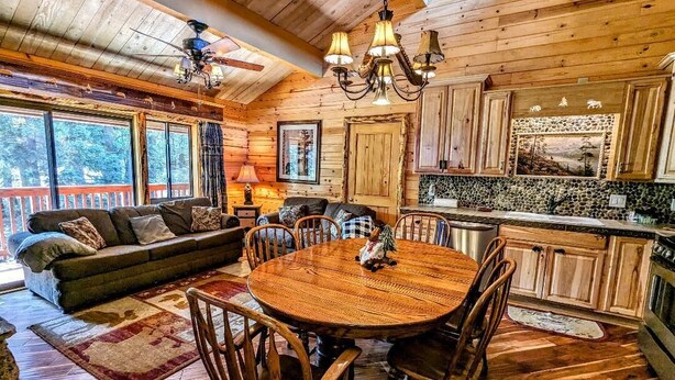 A cozy cabin interior with a wooden dining table surrounded by chairs, a sofa with cushions, a kitchen area with wooden cabinets, and a large window providing a view of the outdoors.