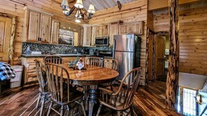 A rustic wooden interior with a round dining table surrounded by chairs, a kitchen area with wooden cabinets and modern appliances, and a chandelier hanging from the ceiling.