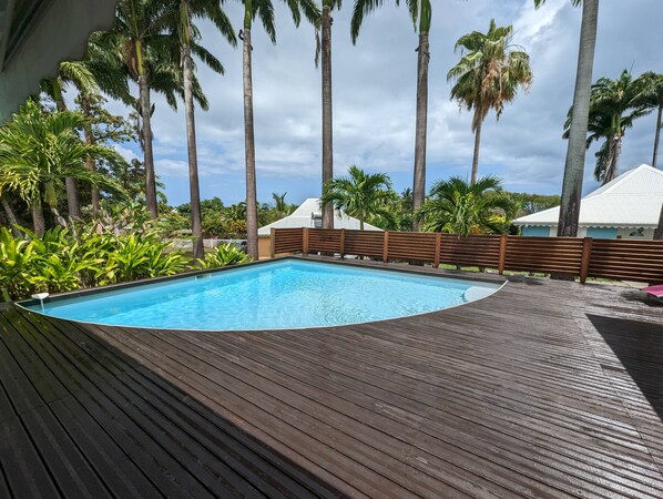 A swimming pool with a wooden deck, surrounded by palm trees and a white building in the background.