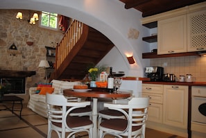 Cozinha acolhedora com lareira de pedra, escada de madeira e área de jantar com cadeiras brancas.