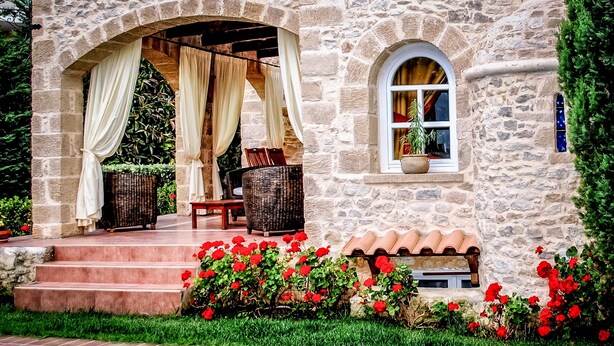 A stone building with a pool, steps leading to an outdoor seating area with wicker furniture, and red flowers.