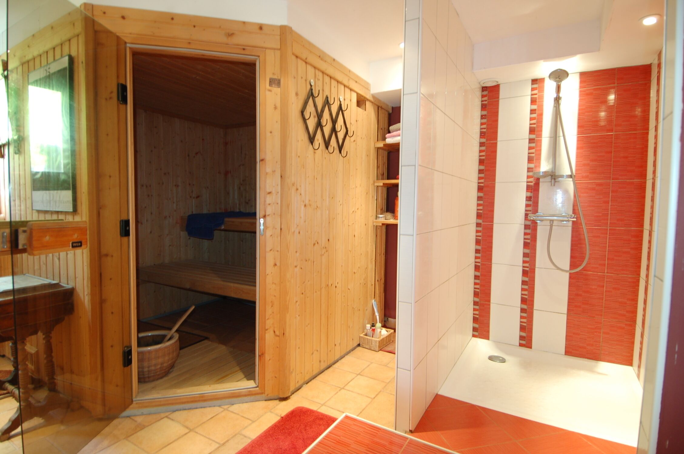 A sauna with a wooden door, a shower area with red tiles, and a small wooden shelf with towels.