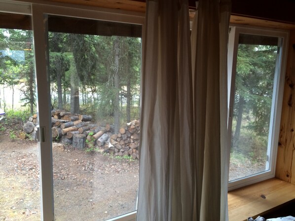 A cabin with a large sliding glass door leading to a forested area with stacked logs. - Log Cabin W Bedroom Loft, Seperate private Bath House and Boat House (Sterling)