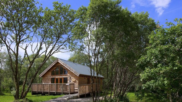 A wooden cabin with a deck, surrounded by trees and a gravel path. - Silver Birch Cabin - Charming Log Cabin with bags of character and hot tub. (Dalavich, Taynuilt)