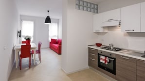 A modern kitchen with white cabinets, a gas stove, and a built-in oven. There is a dining area with a white table and red chairs. A red sofa is visible in the background.