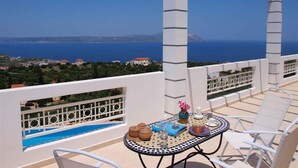 A terrace with a pool, white furniture, and a view of the sea and distant buildings.