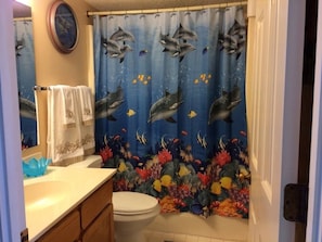 Een badkamer met een douchegordijn met dolfijnenprint, een wit toilet, een houten wastafelmeubel met een blauwe kom, en een spiegel boven de wastafel.
