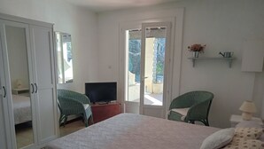 A bedroom with a bed, a television on a stand, two green chairs, a small table, and a shelf with a watering can and flowers. - Vacation (SAINT REMY DE PROVENCE)