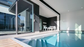 A modern indoor swimming pool with glass walls, a wooden deck, and a barbecue grill outside. - Villa O Bord de l'O Sainte Marine, sea view, swimming pool 29°, beach access (Combrit)