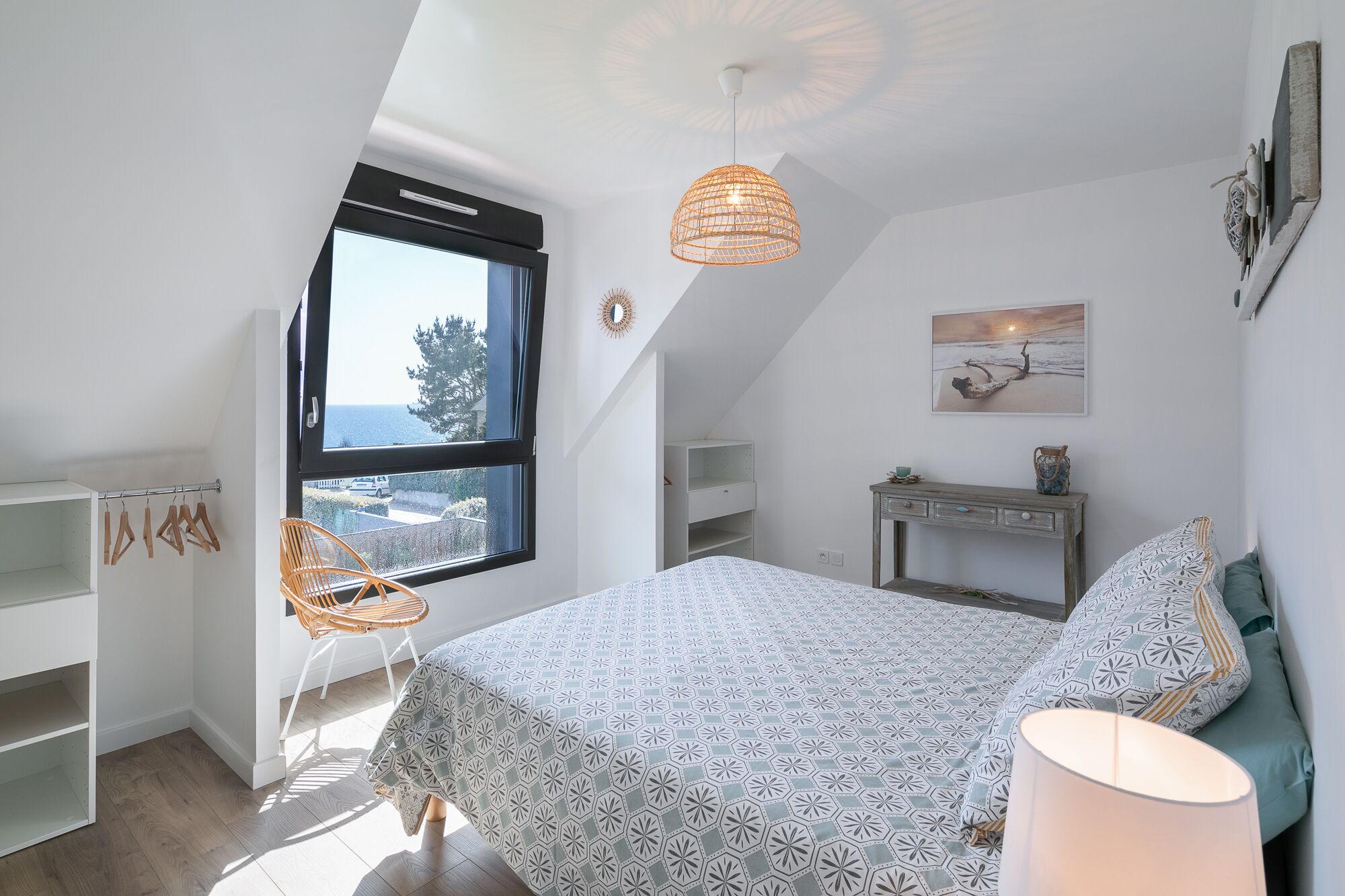 Un dormitorio con cama, escritorio, silla, lámpara colgante y vistas al mar a través de una ventana.