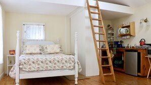 A bedroom with a wooden ladder, a bed with floral bedding, a window with curtains, and a kitchen area with shelves and appliances. - Private Lake at Rippling Waters One Room Cabin (Delhi)