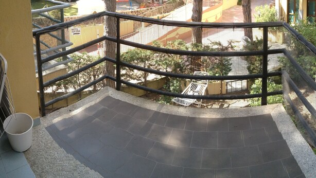 A balcony with a metal railing, a white bucket, and a view of a playground and buildings in the background.