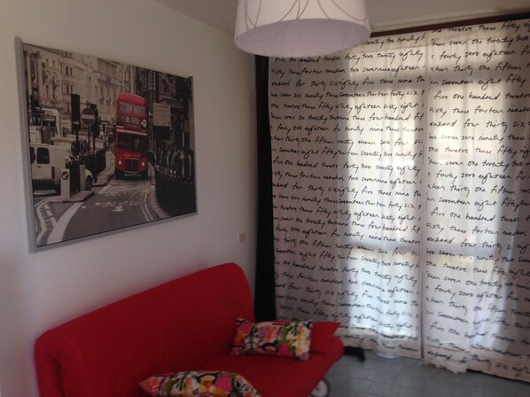 Une pièce avec un canapé rouge, un tableau représentant une scène de rue en noir et blanc, et un rideau orné d’un texte écrit à la main.