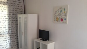 A room with a white wardrobe, a TV on a white stand, and a wall with a colorful drawing.