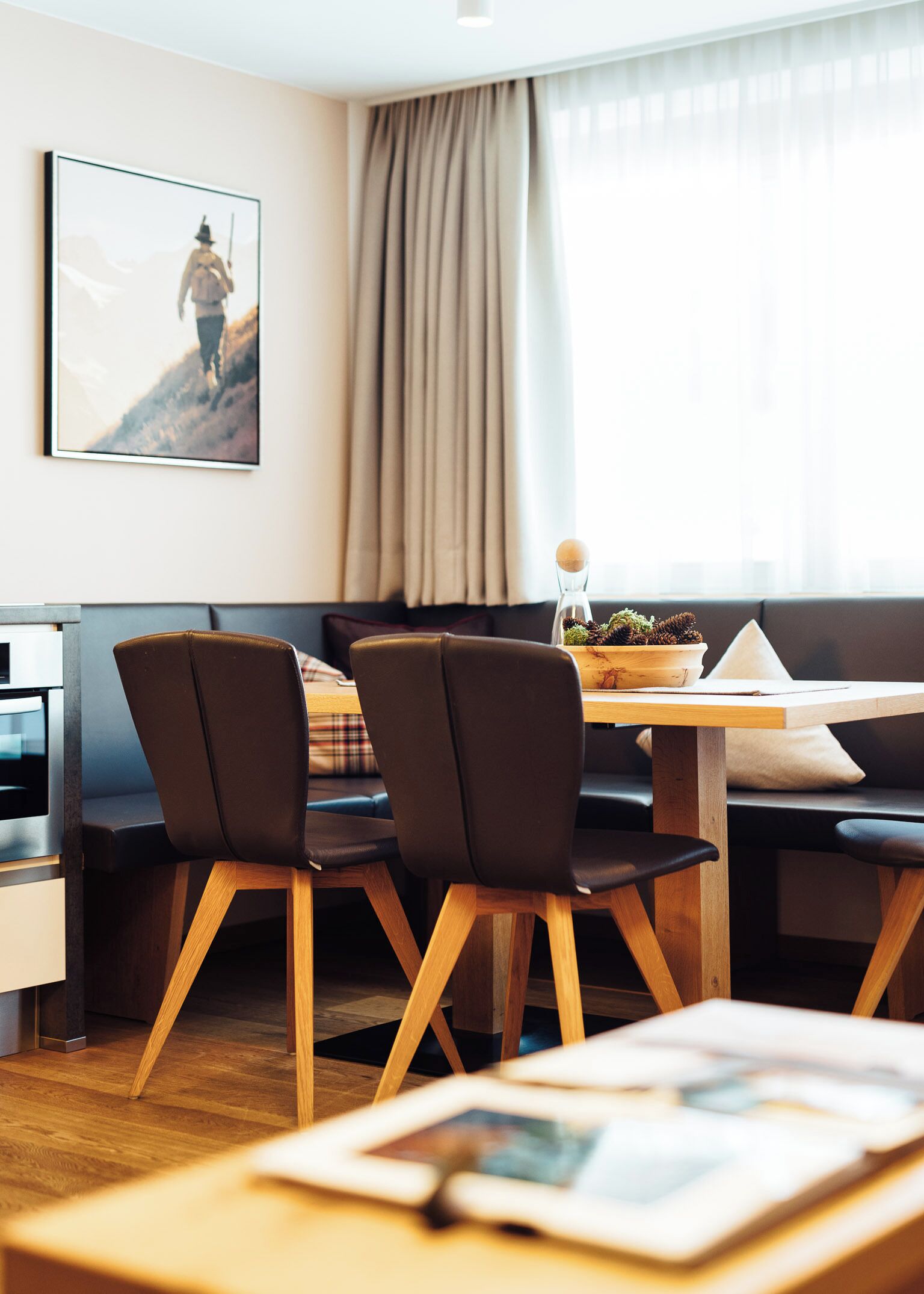 A modern dining area with a wooden table, black chairs, and a bench. A framed picture is on the wall, and a window with curtains is visible.
