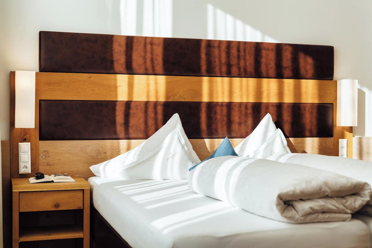 Ein ordentlich bezogenes Bett mit weißen Leinen und einer gefalteten Decke, ein Nachttisch aus Holz mit einem Buch und einer Brille, und ein modernes Kopfstück mit Holz- und dunklen Paneelelementen.