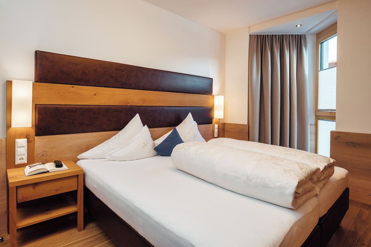 Ein ordentlich gemachtes Bett mit Holz-Kopfstück, ein Nachttisch mit Lampe und ein Fenster mit Vorhängen.