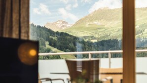 Une pièce avec vue sur un paysage montagneux. On y trouve une table en bois sur laquelle sont posés un vase de fleurs et un objet noir.