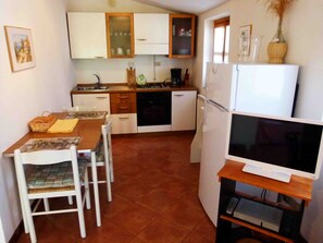 A compact kitchen with white cabinets, a dining table with chairs, a refrigerator, and a television on a wooden stand.