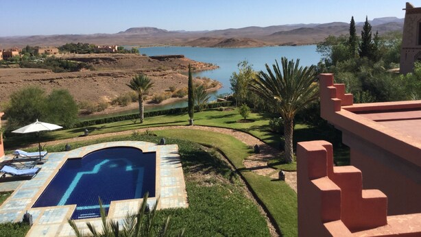 Una piscina con vistas a un lago y montañas, rodeada de vegetación y un edificio con fachada roja.