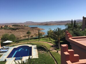 Une piscine avec vue sur un lac et des montagnes, entourée de verdure et bordée d’un bâtiment à la façade rouge.