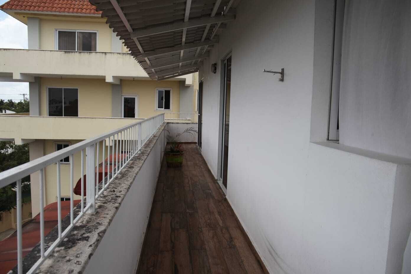 A narrow balcony with a wooden floor, white railing, and a view of adjacent buildings.