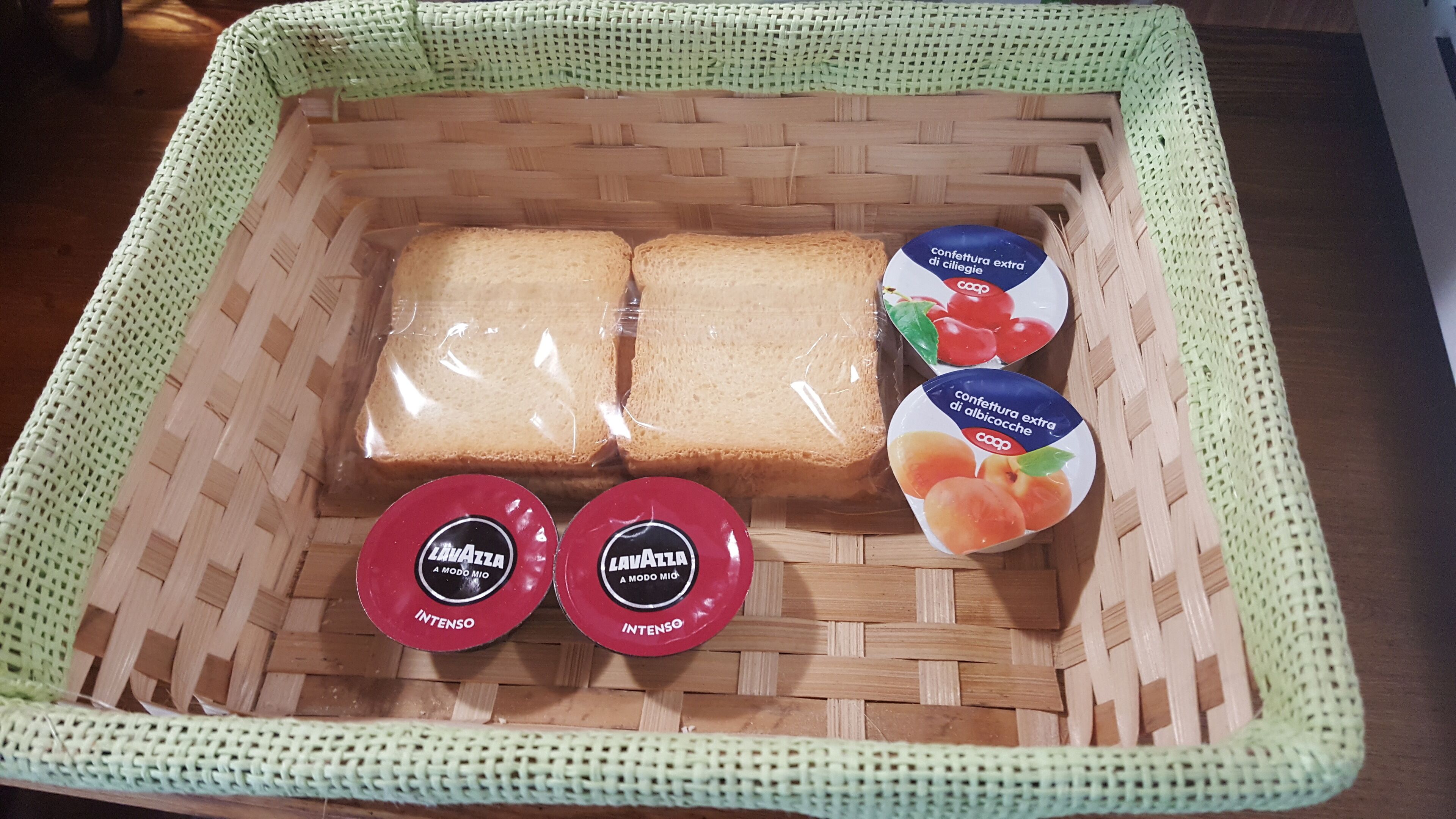 Ein geflochtener Korb mit zwei Brotscheiben, zwei Gläsern Lavazza-Kaffee und zwei Behältern mit extra Käse und Albacoche-Aufstrich.