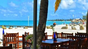 A beachside restaurant with wooden tables and chairs, a view of the ocean, and thatched-roof structures in the background. - Aruba Villa with Pool, just 3 minutes driving from Aruba's best beaches (Noord)