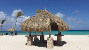 A beach with thatched huts, palm trees, and a clear blue sky. - Aruba Villa with Pool, just 3 minutes driving from Aruba's best beaches (Noord)