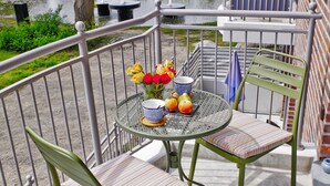Un balcon avec deux chaises, une table ornée d’un vase de fleurs, et une vue sur un canal et des bâtiments en arrière-plan.