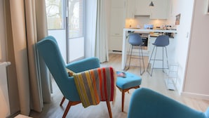 Un salon moderne avec un canapé bleu, une petite table et un plaid à rayures. On aperçoit, au fond, une cuisine avec des meubles blancs et un tabouret de bar.