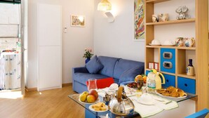 A living room with a blue sofa, a glass table with food and drinks, and shelves with kitchenware.