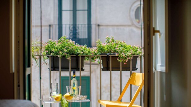 A balcony with two flower planters, a glass table with a bottle and lemons, and a yellow chair.