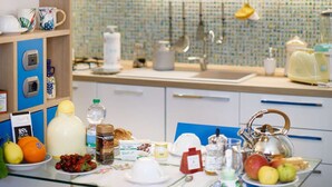 A modern kitchen with wooden shelves, hanging pendant lights, and a glass table set with breakfast items.