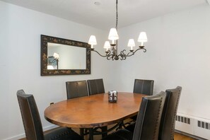 A dining room with a round wooden table, six black upholstered chairs, a chandelier with multiple light fixtures, and a large mirror with a floral frame on the wall. - Villa Valhalla | Unit 2 - 2 Bedroom Platinum (Vail)