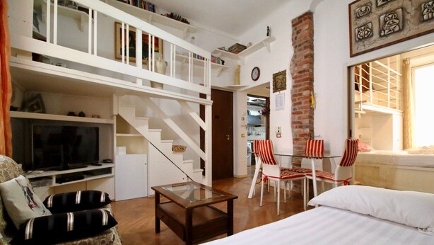 Ein gemütliches Wohnzimmer mit Fernseher, Sofa, Couchtisch und einer Treppe zu einem Erkerraum. Es gibt gestreifte Stühle und eine Ziegelwand.