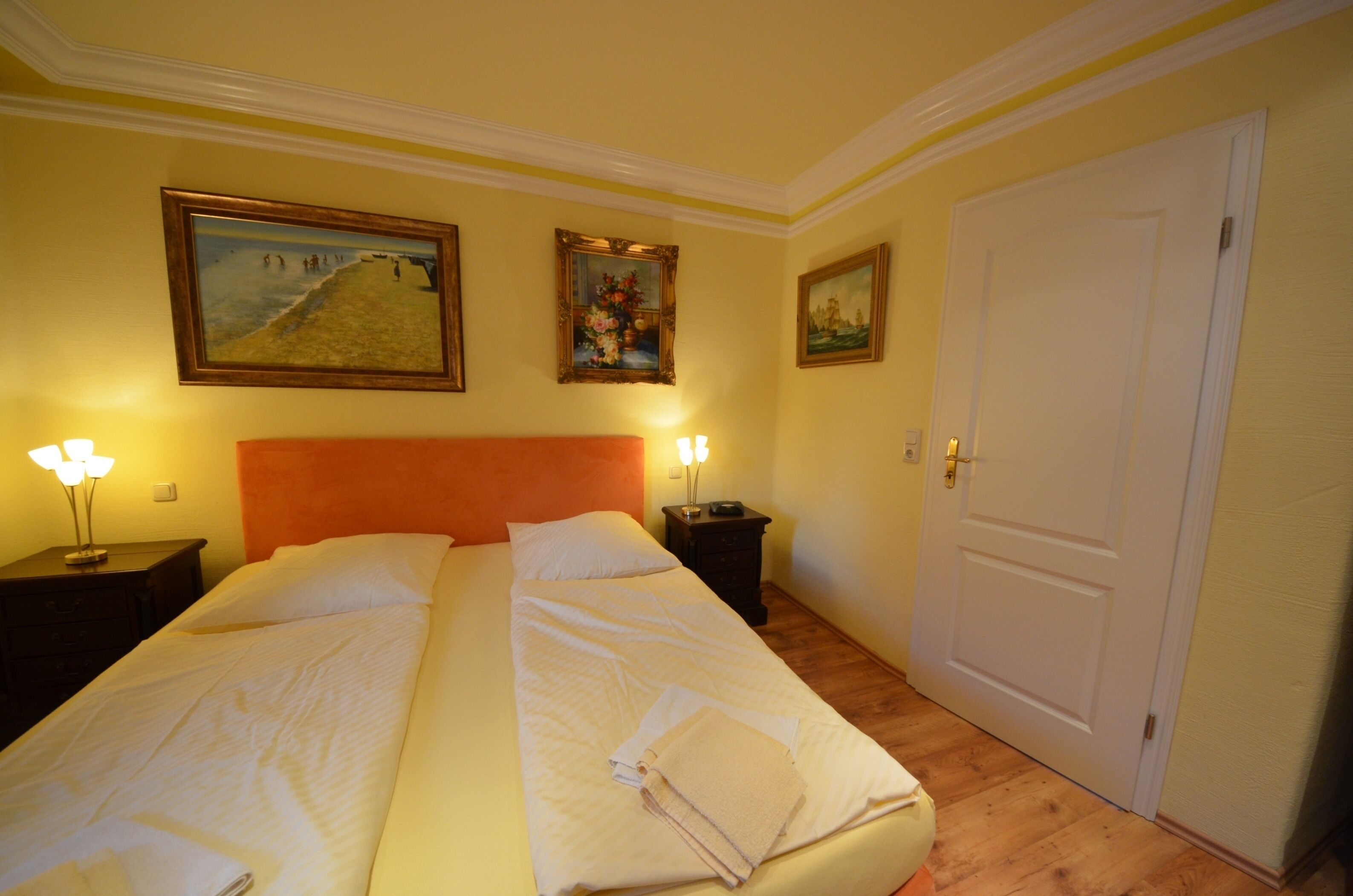 A double bed with white linens, two bedside tables with lamps, and three framed artworks on the wall.
