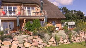 Ein zweigeschossiges Haus mit einem Strohdach, einem Balkon mit roten Stühlen und einer Steinmauer im Vordergrund.