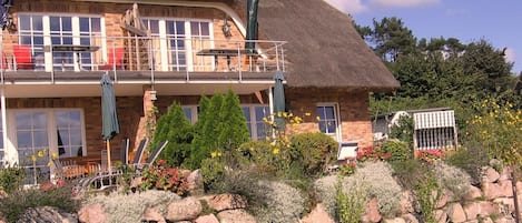 A two-story house with a thatched roof, a balcony with red chairs, and a stone wall in the foreground.