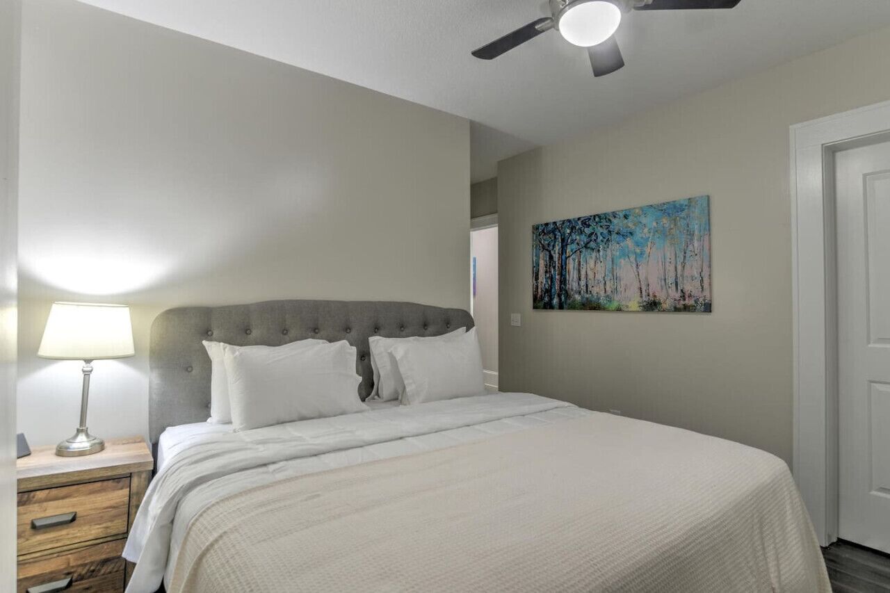 A bedroom with a bed, a nightstand, a lamp, a ceiling fan, and a painting on the wall.