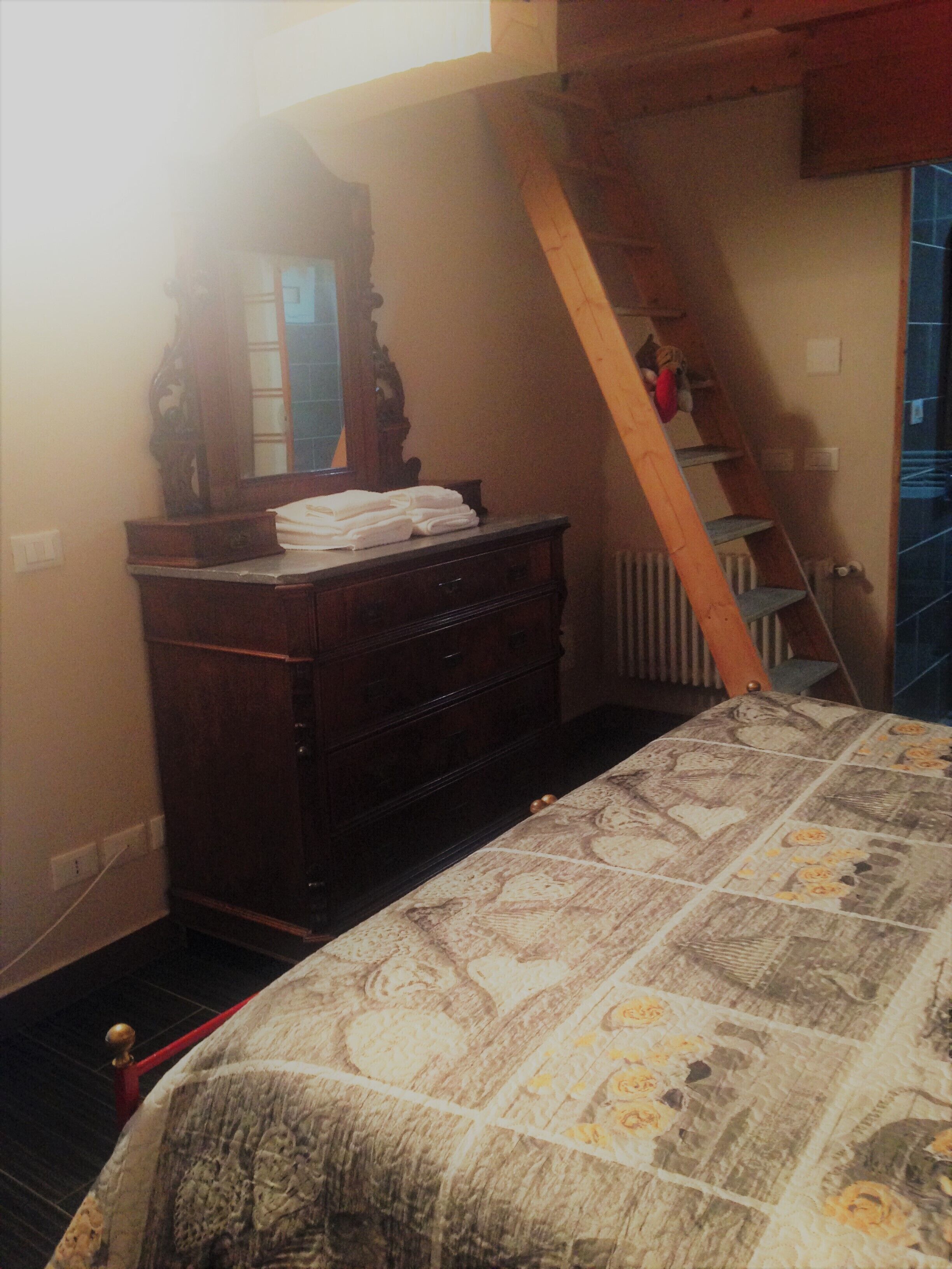 Ein Schlafzimmer mit einem hölzernen Schminktisch, einem Bett mit blumigem Überwurf, einer Holzleiter und einem Fenster mit Blick ins Freie.